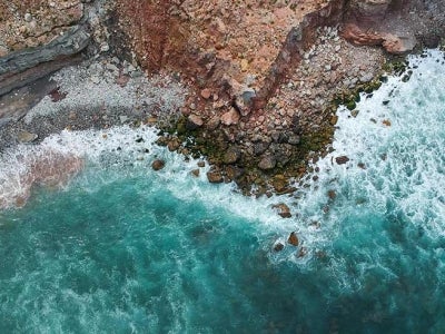 Aerial top view of sea waves hitting rocks on the beach with turquoise sea water.