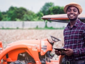 African farmer is using a tablet on the background of working tractor with a cultivator in the field