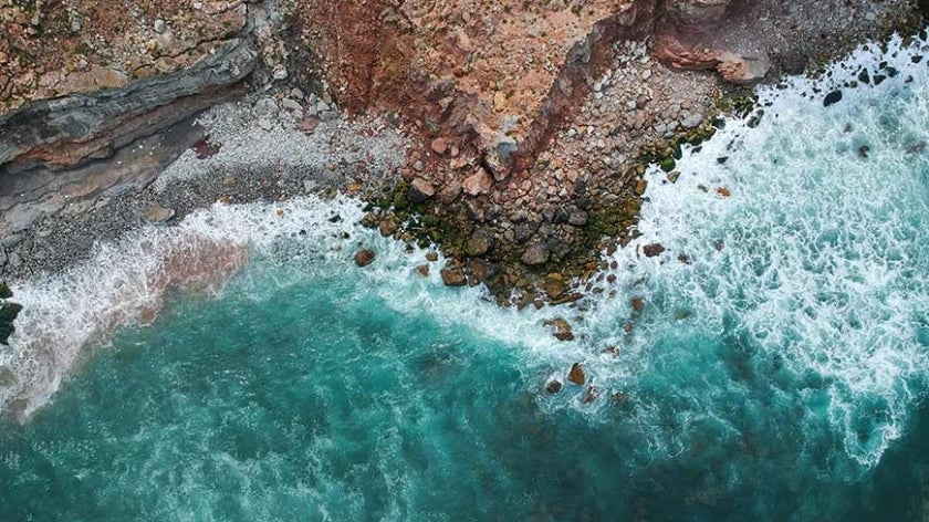 Aerial top view of sea waves hitting rocks on the beach with turquoise sea water.