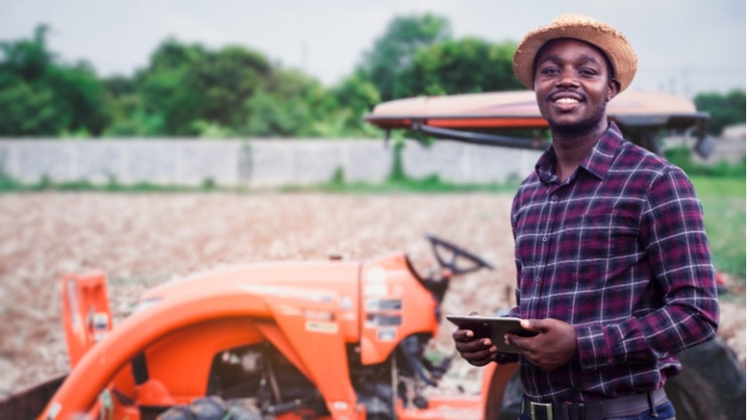 African farmer is using a tablet on the background of working tractor with a cultivator in the field